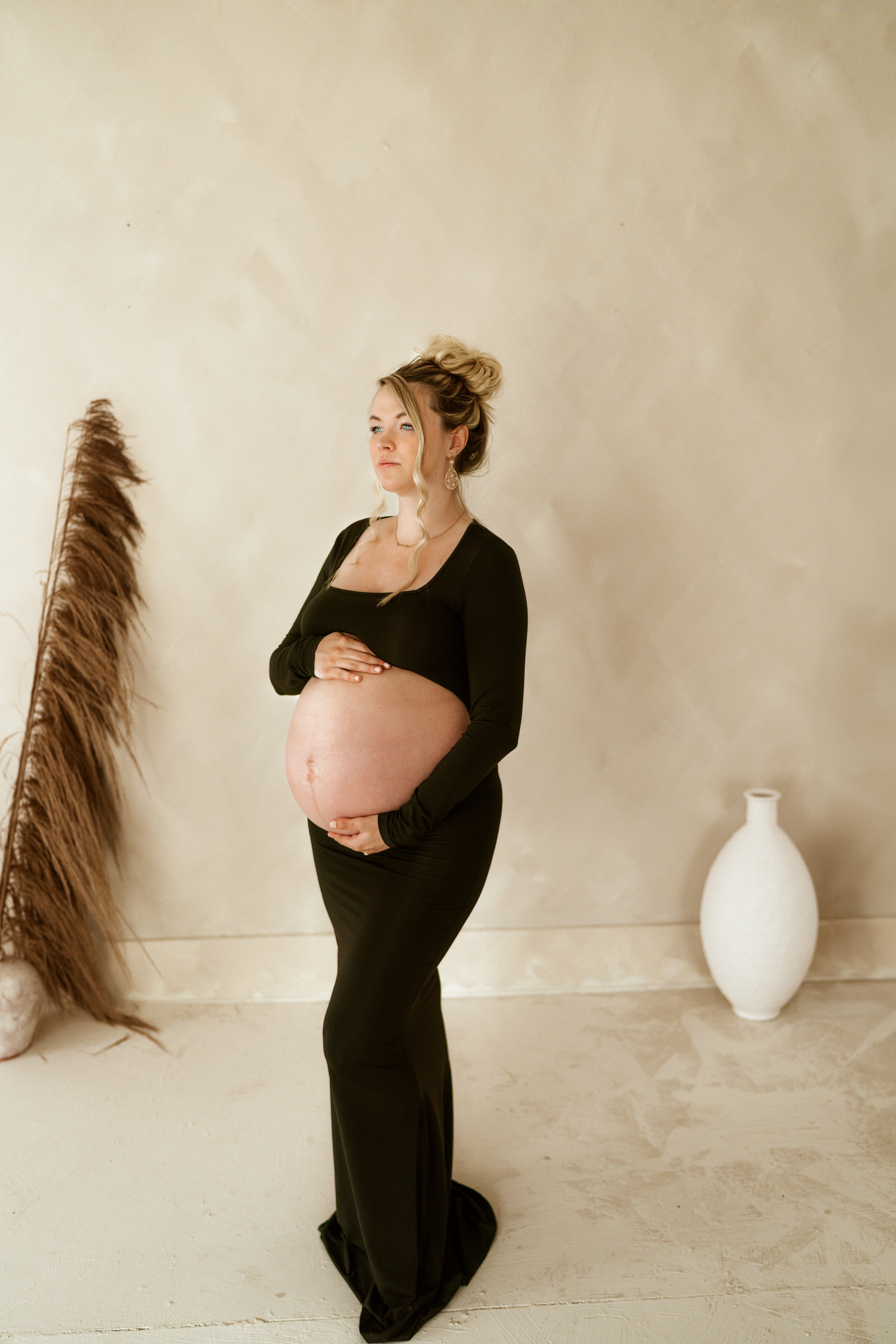 A pregnant woman in a black dress cradles her baby bump, standing gracefully in Maria Angeles Photography studio in Erie, Pa